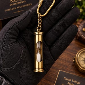 May include: A brass hourglass keyring featuring a cylindrical metal frame with a clear glass centre containing white sand. The hourglass is attached to a brass chain and a carabiner-style clip, designed for portability and functional timekeeping.