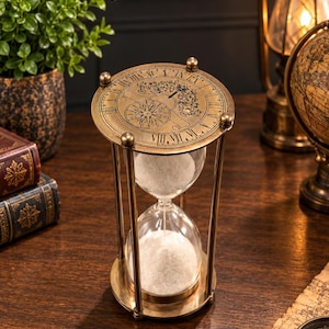 Puede incluir: Un reloj de arena de estilo antiguo con marco de latón y arena blanca. La placa circular superior presenta números romanos grabados, una rosa de los vientos y motivos florales. Los bulbos de vidrio están sujetos por cuatro pilares verticales de latón, conectando las bases circulares superior e inferior en un diseño clásico y funcional.