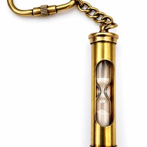 May include: A brass keyring featuring a cylindrical hourglass with white sand, connected by a short metal chain to a screw-lock carabiner. The metallic finish has a warm, golden tone, and the glass centre reveals the fine sand inside.