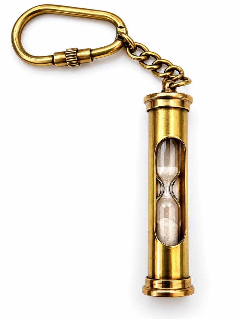 May include: A brass keyring featuring a cylindrical hourglass with white sand, connected by a short metal chain to a screw-lock carabiner. The metallic finish has a warm, golden tone, and the glass centre reveals the fine sand inside.
