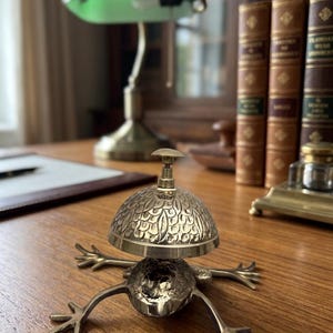 Könnte beinhalten: Eine Schreibtischklingel aus Messing in Froschform mit einem strukturierten, blattartigen Muster auf der Kuppel. Die Klingel verfügt über einen kleinen Knopf an der Oberseite und vier ausgestreckte Beine aus poliertem Metall mit detailliertem Design.