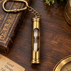 May include: A brass hourglass keyring featuring a cylindrical metal frame with a central glass window revealing white sand. The hourglass is attached to a brass oval carabiner by a short metal chain, creating a functional accessory.