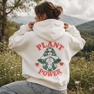 Könnte beinhalten: Ein cremefarbener Kapuzenpullover mit einem Grafikdruck auf der Rückseite. Das Motiv zeigt eine Person mit lockigem Haar und roter Sonnenbrille, die auf einer Kuh sitzt, umrahmt vom roten Schriftzug "PLANT POWER" und zwei kleinen roten Herzen.