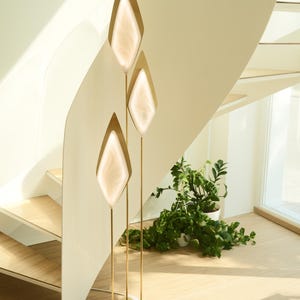 May include: A floor lamp featuring three slender gold-toned metal stems rising from a triangular base. Each stem supports a diamond-shaped frame containing a translucent, glowing white panel with a soft, swirling internal texture.