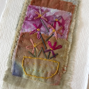 May include: Embroidered textile art with a floral design. The artwork features a yellow pot with pink and yellow flowers, set against patches of brown, purple, and blue fabric. The edges of the fabric are frayed, giving a rustic look.