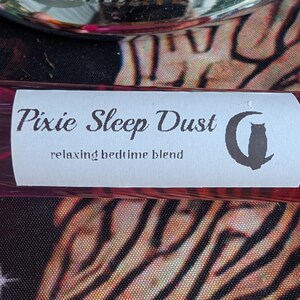 May include: A cylindrical glass bottle with a dark red, striped pattern. A white rectangular label is affixed to the front, featuring the black text Pixie Sleep Dust, the phrase relaxing bedtime blend, and a black silhouette of a cat.