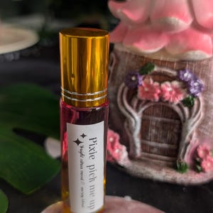 May include: A cylindrical glass roller bottle with a shiny gold cap and a pink liquid inside. A white rectangular label on the front features black text that reads Pixie pick me up, bright citrus mood & energy blend.