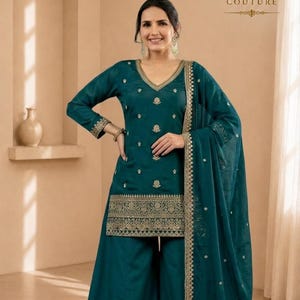 Könnte beinhalten: Ein petrolfarbenes Salwar-Kameez-Set bestehend aus einer langärmeligen Tunika mit Goldstickerei, V-Ausschnitt und einer weiten Hose. Ein passender transparenter Dupatta mit goldener Bordüre und goldenen Motiven ergänzt das Set. Text: KC KYORA COUTURE.