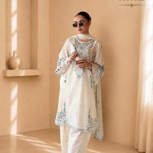 May include: An off-white long-sleeved tunic and matching trousers set featuring intricate blue embroidery along the neckline, cuffs, and hem. The outfit includes a coordinating sheer dupatta draped over the shoulders, paired with white strappy sandals.