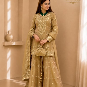 May include: A gold-toned ensemble featuring a long-sleeved tunic and wide-leg trousers, both adorned with intricate gold embroidery. The outfit includes a matching sheer dupatta and a small drawstring pouch. The text KC KYORA COUTURE is visible.