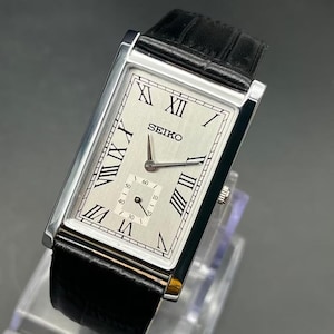 May include: A rectangular Seiko wristwatch with a silver dial, black Roman numeral hour markers, and a sub-dial for seconds. The watch features silver-tone hands and a black leather strap with a textured finish, secured by a polished silver case.
