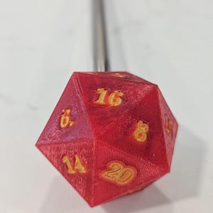 May include: A red twenty-sided die with yellow numbers, featuring a metallic rod extending from the top. The die displays visible layer lines from a 3D printing process, with the number 20 clearly visible on the front-most triangular face.