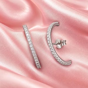 May include: A pair of silver-toned ear climbers featuring a curved bar design encrusted with small, clear, sparkling stones. One earring is positioned vertically, while the other is shown from the back, revealing the post and butterfly clutch backing.