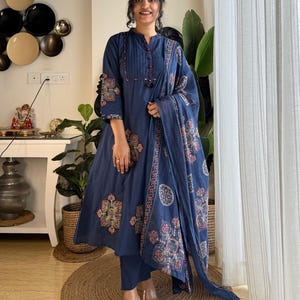 Può includere: Un completo a tre pezzi blu navy composto da un kurta a maniche lunghe con corpetto plissettato e bottoni, pantaloni abbinati e un dupatta coordinato. Il tessuto presenta un motivo floreale ripetitivo in tonalità rosso e crema tenui.