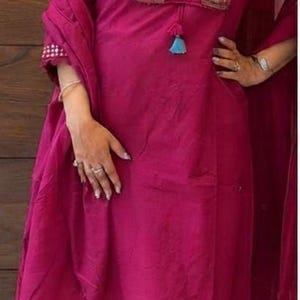 May include: A vibrant magenta long-sleeved tunic and matching wide-leg trousers set. The tunic features a V-neckline with intricate embroidery, small circular mirrors, and a blue tassel detail. The ensemble includes a coordinating magenta dupatta.