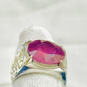Könnte beinhalten: Ein Silberring mit einem großen, oval geschliffenen, tief rosa-roten Edelstein in einer Krappenfassung. Die Ringschiene zeigt an den Seiten ein kompliziertes, durchbrochenes Filigranmuster, das eine detaillierte metallische Struktur bildet.