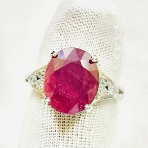 Könnte beinhalten: Ein Silberring mit einem großen, oval geschliffenen, tief rosa-roten Edelstein in einer Vier-Krappen-Fassung. Die Ringschiene ist mit kleinen, klaren, rund geschliffenen Steinen besetzt, die beidseitig des zentralen Edelsteins eingelassen sind.