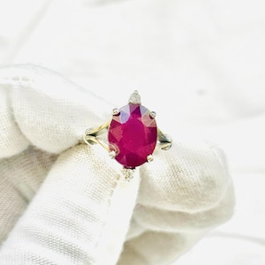 Könnte beinhalten: Ein silberfarbener Ring mit einem großen, oval geschliffenen, tief rosa-roten Edelstein in einer Krappenfassung. Die Ringschiene ist gespalten und an der Ober- und Unterseite des zentralen Edelsteins befinden sich kleine, klare, rund geschliffene Akzentsteine.