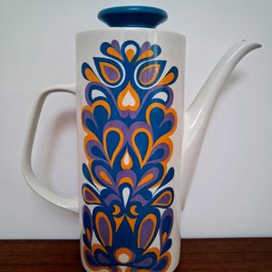 May include: A tall white ceramic coffee pot featuring a cylindrical body, a curved handle, and a long spout. The exterior is decorated with a repeating vertical pattern of stylized teardrop shapes in vibrant shades of blue, orange, and purple.