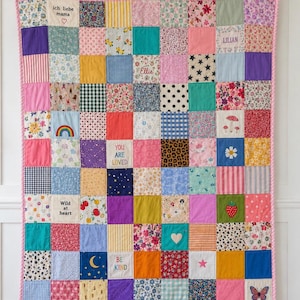 Puede incluir: Una colcha de retazos con una cuadrícula de cuadrados de tela coloridos con varios patrones que incluyen flores, rayas, lunares y estampados de animales. Varios cuadrados contienen texto bordado como ich liebe mama, LILIAN, Ellie, YOU ARE LOVED, Wild at heart y BE KIND.