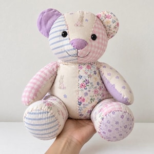 Könnte beinhalten: Ein Patchwork-Teddybär aus verschiedenen Stoffmustern, darunter hellblaue Streifen, rosa Vichy-Karo, Blumenmuster und einfarbiges Lavendel. Der Bär hat schwarze Knopfaugen, eine lavendelfarbene Nase und kleine Hasenillustrationen.