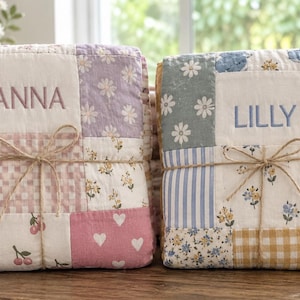 Könnte beinhalten: Zwei gefaltete Patchwork-Steppdecken, die mit Bindfaden zusammengebunden sind. Die linke Decke zeigt rosa-weiße Blumen-, Kirsch-, Herz- und Karomuster mit dem eingestickten Namen ANNA. Die rechte Decke zeigt blau-gelb-weiße Blumen-, Stern- und Walmuster mit dem eingestickten Namen LILLY.