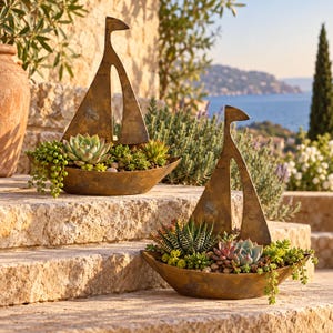 May include: Two bronze-colored metal sailboat-shaped planters, each containing a variety of small green and reddish succulents. The planters feature a tall, stylized sail silhouette and a curved hull base, filled with soil and small pebbles.
