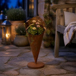 May include: A tall, cone-shaped metal planter with a rusted, weathered finish, standing on a circular base. The planter is filled with a variety of small, blooming flowers in shades of white and pink, with green foliage spilling over the edges.