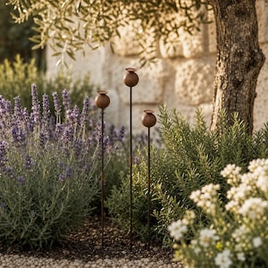 May include: Three metal garden stakes featuring spherical poppy seed pod toppers in a rusted brown finish. Each topper is mounted on a thin, straight vertical rod, creating a slender silhouette that stands upright among lavender and green foliage.