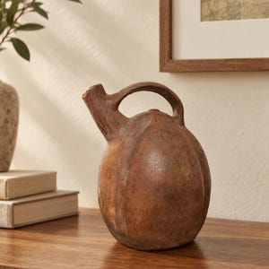 Puede incluir: Un recipiente de cerámica de color terracota, de forma redonda con una superficie texturizada y desgastada. El objeto presenta un único pico inclinado y un asa curva que conecta la parte superior del pico con el cuerpo principal del recipiente.