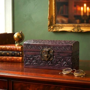 May include: A rectangular dark purple wooden storage box with a domed lid, featuring an intricate embossed floral pattern and a central brass-colored metal latch. The box is positioned on a wooden surface next to a pair of gold-rimmed spectacles.