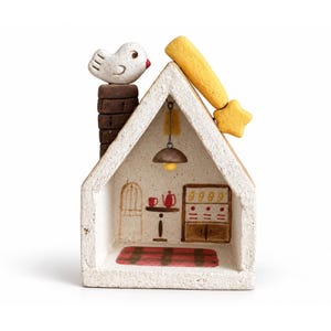 May include: A small, off-white ceramic house figurine with a textured finish. A white bird sits on a brown chimney. A yellow shooting star is attached to the roof. Inside, a painted scene shows a table with a red teapot, a cupboard, and a red rug.