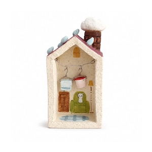 May include: A miniature house figurine with a textured cream exterior, a reddish-brown roof with blue accents, and a brown chimney. The interior features a painted green armchair, a brown cabinet, a blue rug, and two small buckets hanging from a wire.