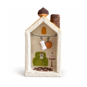 May include: A small, off-white, house-shaped ceramic sculpture with a brown chimney and a dark brown roof accent. Inside, a wire holds a white bone and an orange ball. The back wall features a painted green armchair, a dog, and a cake.