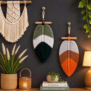 Puede incluir: Dos tapices de macramé en forma de pluma con patrones de colores en capas. Una pluma muestra verde oliva, blanco y verde oscuro, mientras que la otra presenta gris claro, marrón y naranja. Ambas cuelgan de varillas de madera con cuentas oscuras.