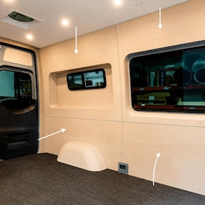 Op de afbeelding: Het interieur van een bestelwagen met lichtgekleurde houten panelen op de wanden, een donkergrijze vloerbedekking en twee ramen met zwarte kozijnen. De wanden bevatten uitsparingen en witte pijlen die naar de montagepunten van de panelen wijzen.