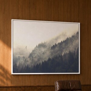 May include: A rectangular framed wall art print featuring a misty forest landscape with dark evergreen trees and fog, mounted on a wood-paneled wall above an orange desk, a brown leather office chair, and two green textured stools.