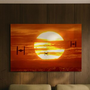 May include: A rectangular wall art print featuring a vibrant orange and yellow sunset with several dark TIE fighter silhouettes flying across the sky. The scene depicts a large, bright sun partially obscured by horizontal clouds above a dark horizon.