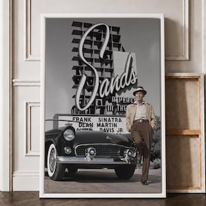 May include: A framed black and white wall art print featuring a vintage car parked in front of the Sands hotel sign. The sign displays the text Sands, A Place In The Sun, Frank Sinatra, Dean Martin, and Davis Jr. A person stands beside the car.