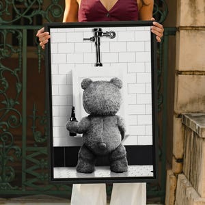 May include: A framed wall art print featuring a black and white image of a fuzzy teddy bear standing at a white porcelain urinal. The bear is holding a dark glass beer bottle in one hand. The background shows white subway tiles and a chrome flush valve.
