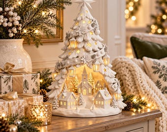Puede incluir: Una escultura de árbol de Navidad de cerámica blanca con un centro hueco que revela un pueblo en miniatura con una iglesia y casas. El árbol está decorado con recortes de estrellas, linternas colgantes y una estrella en la punta.