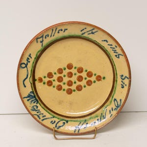 May include: A round, antique ceramic plate with a cream-colored center and a brown rim. The plate features a diamond-shaped pattern of brown and green dots in the center and blue script around the edge.