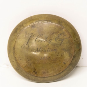 May include: A round, weathered brass snuff box with a hinged lid. The surface features an engraved inscription that reads J. Cooley Esq., Moulton, 1811. The metal has a dull, aged patina with visible surface marks and a distinct circular border.