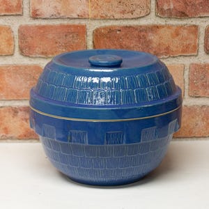 May include: A blue ceramic lidded container with a textured, geometric pattern. The container is round with a lid and a small knob on top. The background is a brick wall.