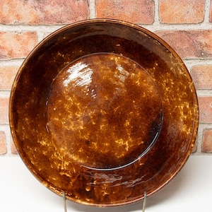 May include: A round, brown ceramic bowl with a speckled glaze. The bowl is displayed on a metal stand against a brick wall background. The bowl's interior has a glossy finish.