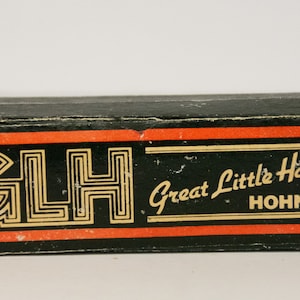 May include: A black box with a red and gold label. The label says "GLH Great Little Harp HOHNER".