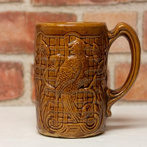May include: Brown ceramic mug with a detailed relief of a bird and foliage. The mug has a large handle and a textured surface. The design is set against a grid pattern. A decorative piece for beverages.