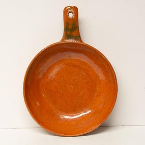 May include: A handmade, orange ceramic pan with a handle. The pan has a round, shallow bowl and a handle with a hole for hanging. The handle has a green and brown color gradient. The pan is likely for cooking.