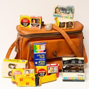 May include: A collection of vintage photography equipment, including film boxes, flashbulbs, and Magicubes, arranged on and around a brown leather camera bag. The film boxes feature colorful graphics and the text "Triple Print Color Film".