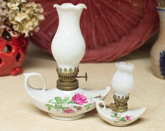 May include: Two white porcelain oil lamps with floral designs. The larger lamp has a handle and a white glass chimney. The smaller lamp is a miniature version of the larger lamp.